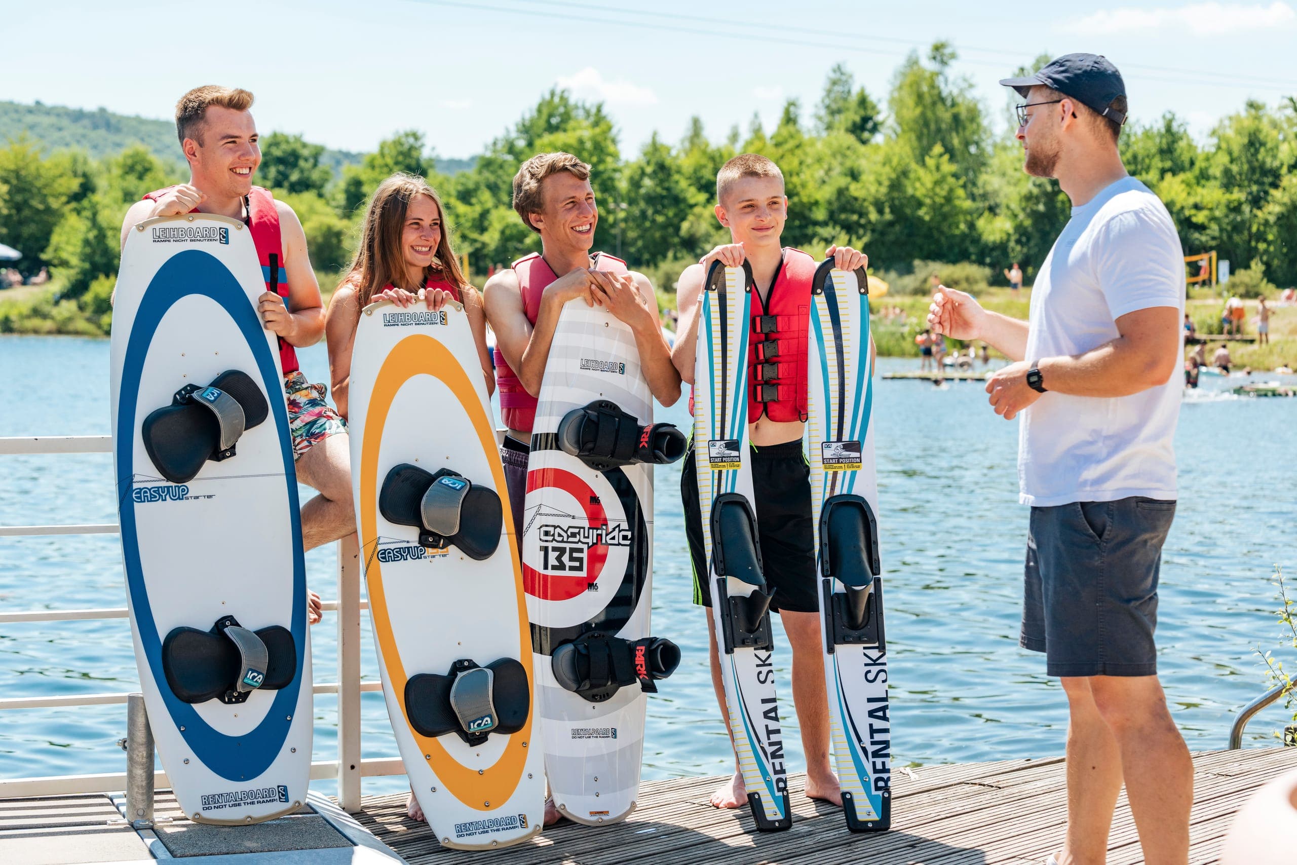 Trainer erklärt vier Jugendlichen in Schwimmwesten mit Wakeboards und Wasserskiern am Steg den Start