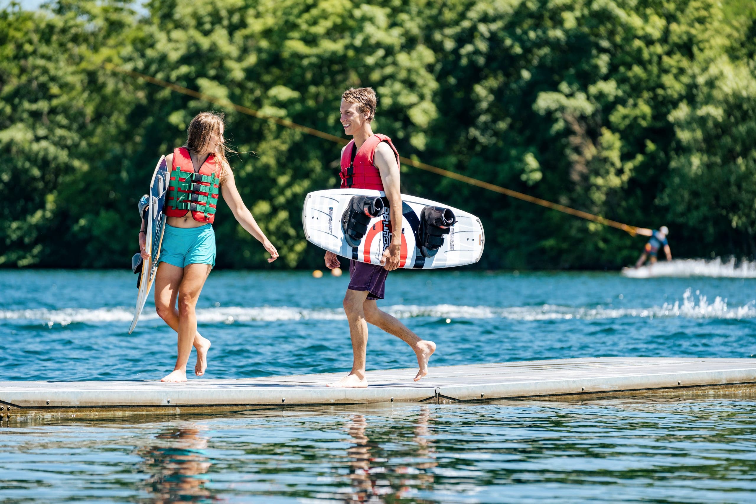 Zwei Jugendliche in Schwimmwesten tragen ihre Wakeboards barfuß über den Schwimmsteg