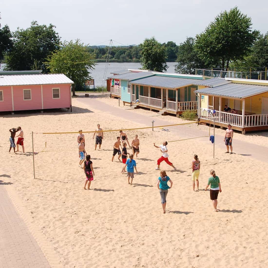 Jugendliche spielen Beachvolleyball vor den bunten Holzhütten am See