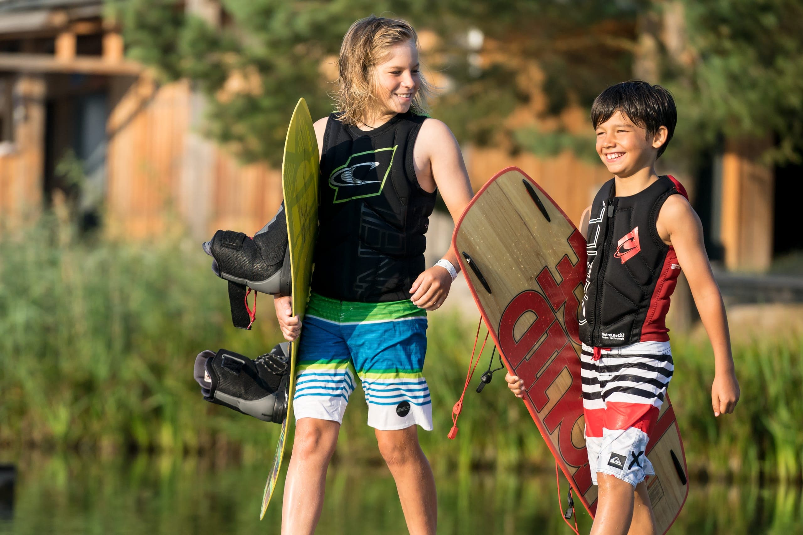 Zwei Jungen in Prallschutzwesten lachen mit ihren Wakeboards am Ufer