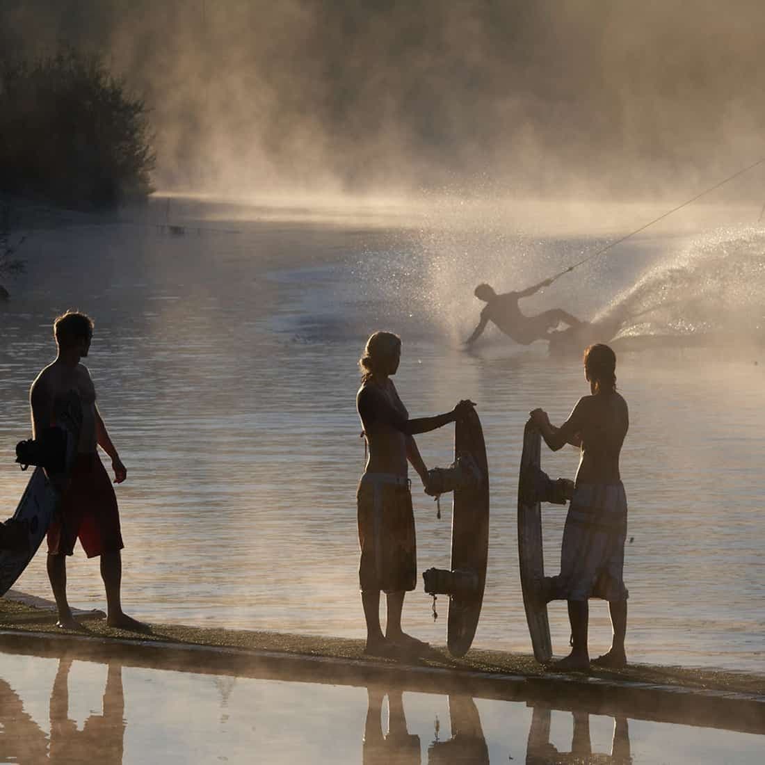 Wakeboarder mit Boards am Steg im Gegenlicht, dahinter eine Fahrt durch Nebel über dem See