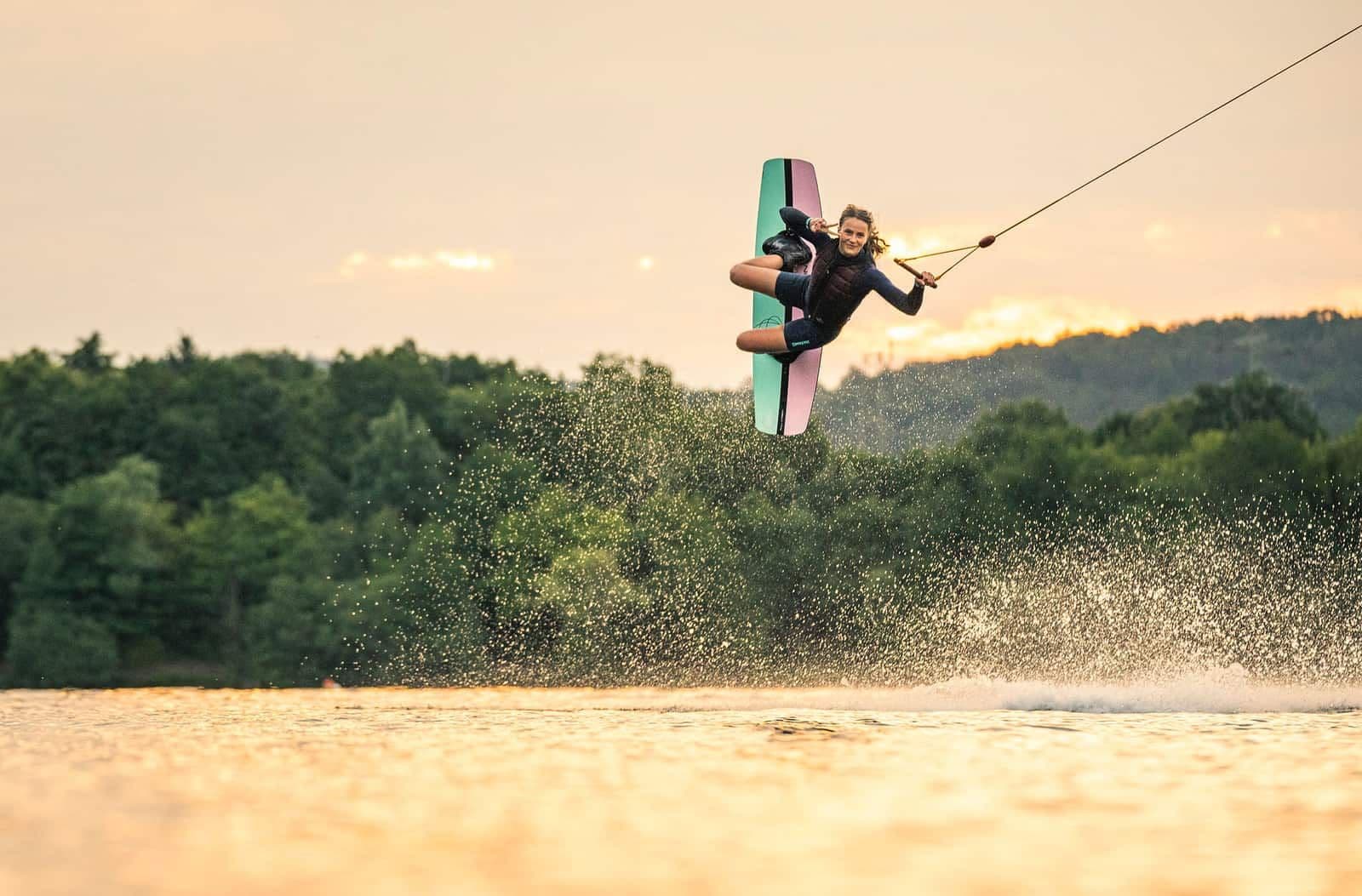 Wakeboarderin mit Weste in der Luft, dahinter Bäume im Abendlicht