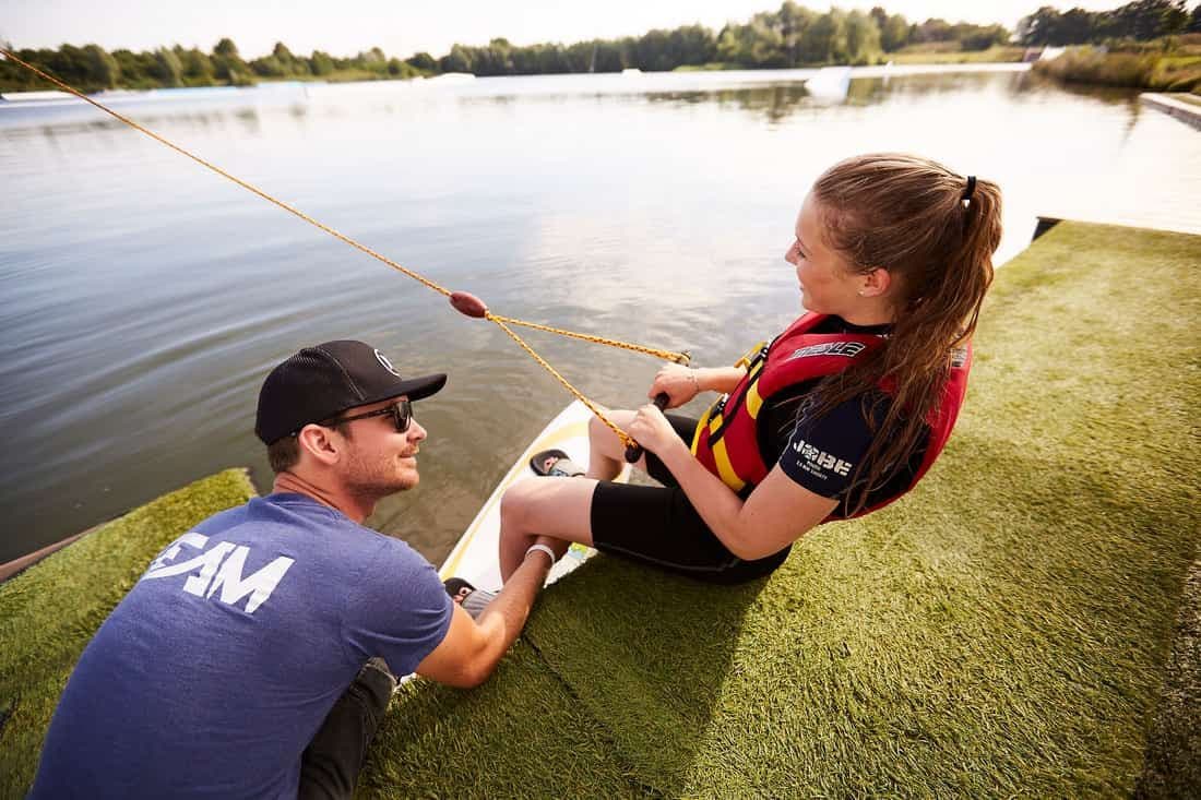 Ein Trainer erklärt einer Wakeboarderin mit Weste den Start vom Steg